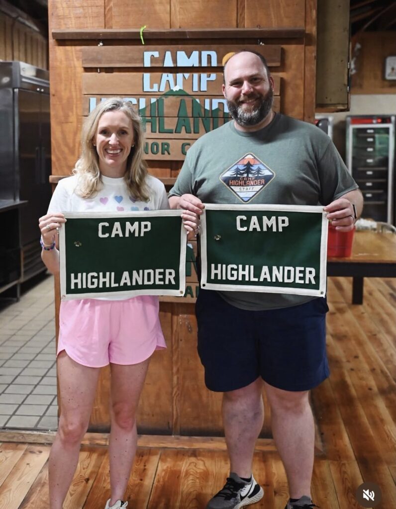 Smiling woman and man in a camp dining hall each holding a green Camp Highlander felt banner