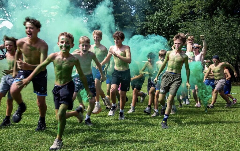 Campers with green face paint running across a camp field through green color smoke