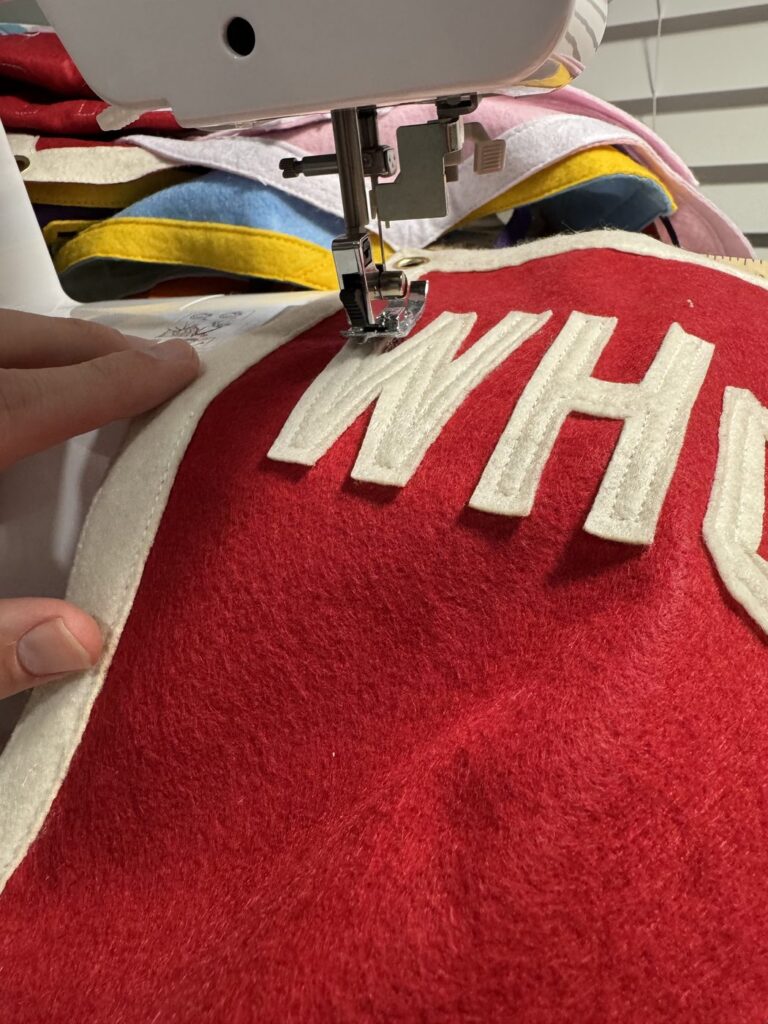 Felt lettering being stitched onto a red custom camp pennant at a sewing machine with colorful felt behind it