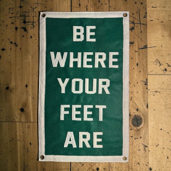 Tall green felt banner reading Be Where Your Feet Are hanging on a wood wall