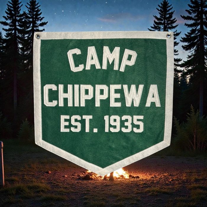 Green Camp Chippewa Est. 1935 felt banner in front of pine trees and a glowing campfire at night