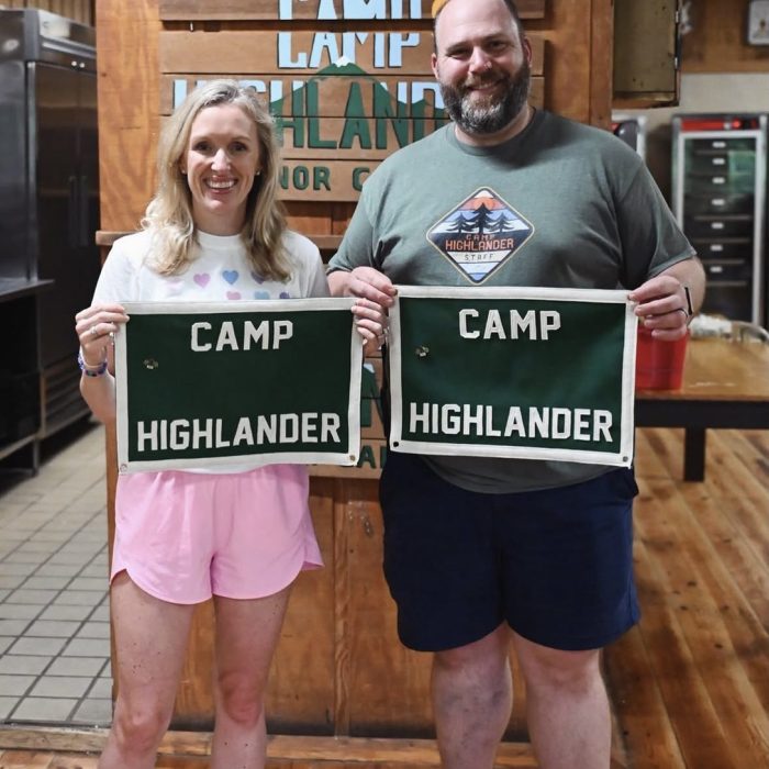 Smiling woman and man in a camp dining hall each holding a green Camp Highlander felt banner