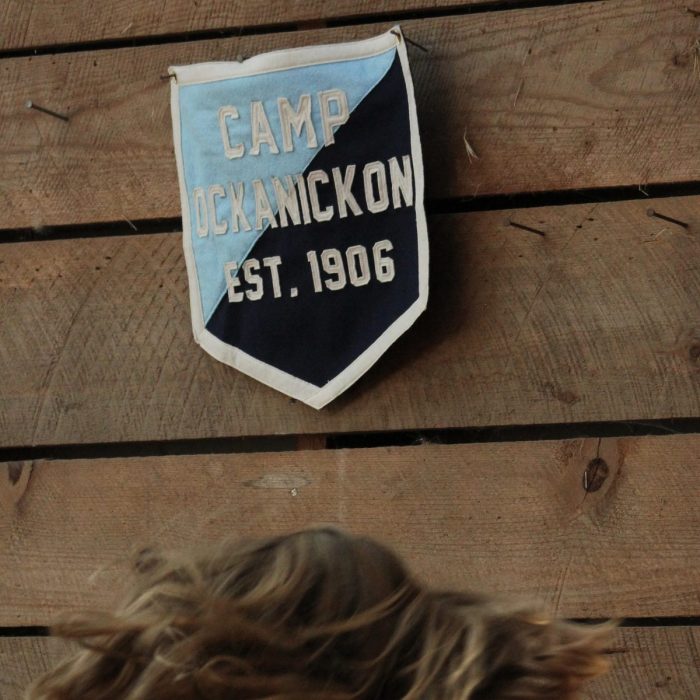 Small felt Camp Ockanickon Est. 1906 badge on a rustic wood cabin wall with a camper’s windswept hair in the foreground