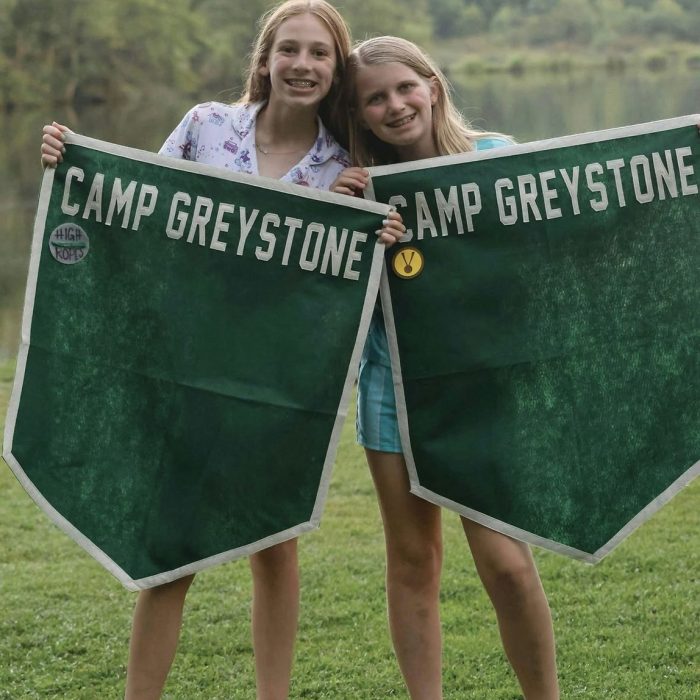 Two smiling campers standing on a lawn holding green Camp Greystone felt banners