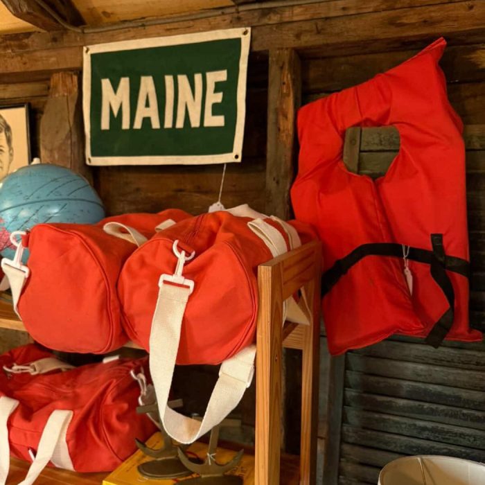 Green felt Maine sign hanging above orange life jackets and duffel bags in a rustic camp boathouse